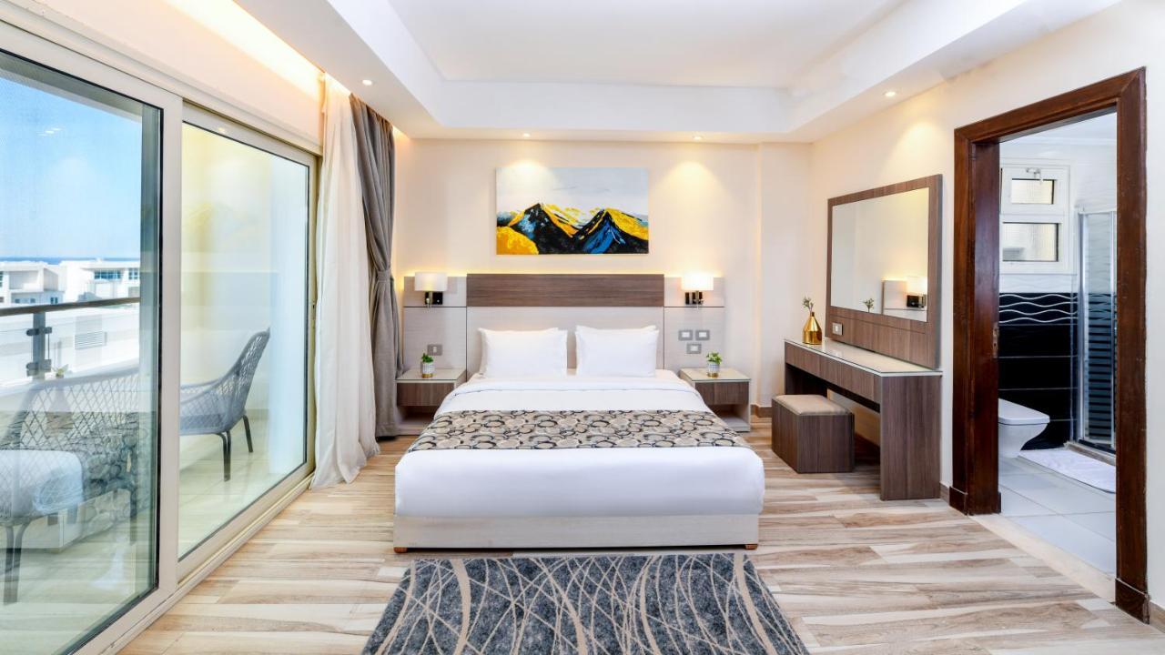 Tolip Resort El Galala Majestic Ain Suchna Exterior foto The photo shows a modern hotel room featuring a large bed with white linens and a decorative throw. The room has a neutral color palette, accented with warm lighting. There is a piece of artwork above the bed depicting mountains. To the left, there a