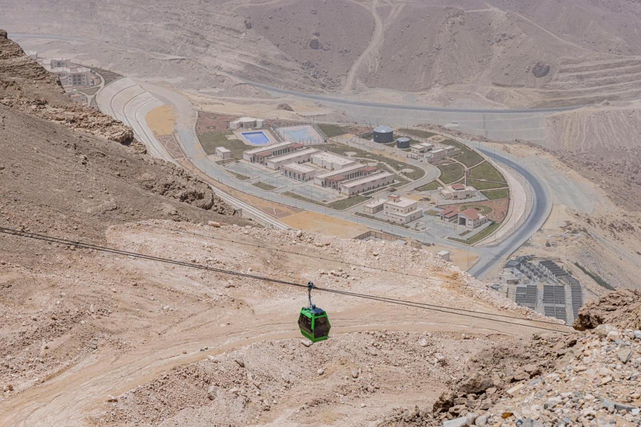 Tolip Resort El Galala Majestic Ain Suchna Exterior foto The photo shows a mountainous landscape with a steep, dry terrain. In the foreground, there is a green cable car or gondola traveling along a cable. Below, a complex of buildings and roads can be seen, likely part of a resort or recreational area. Th