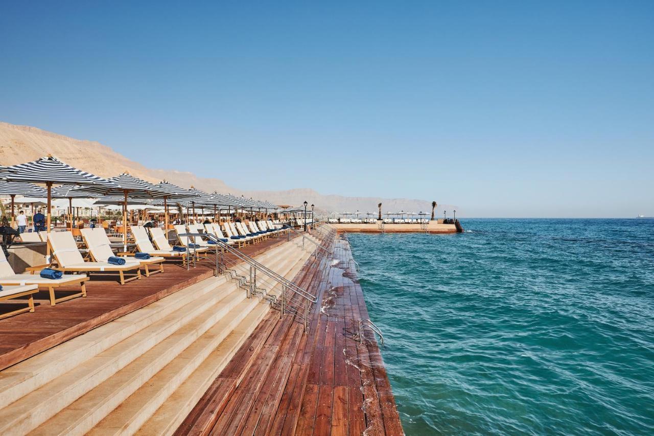 Tolip Resort El Galala Majestic Ain Suchna Exterior foto The photo shows a seaside area featuring a wooden deck that extends into the water. On the left side, there are several lounge chairs arranged neatly under shade structures, suggesting a relaxing atmosphere. The ocean is visible in the foreground, wi