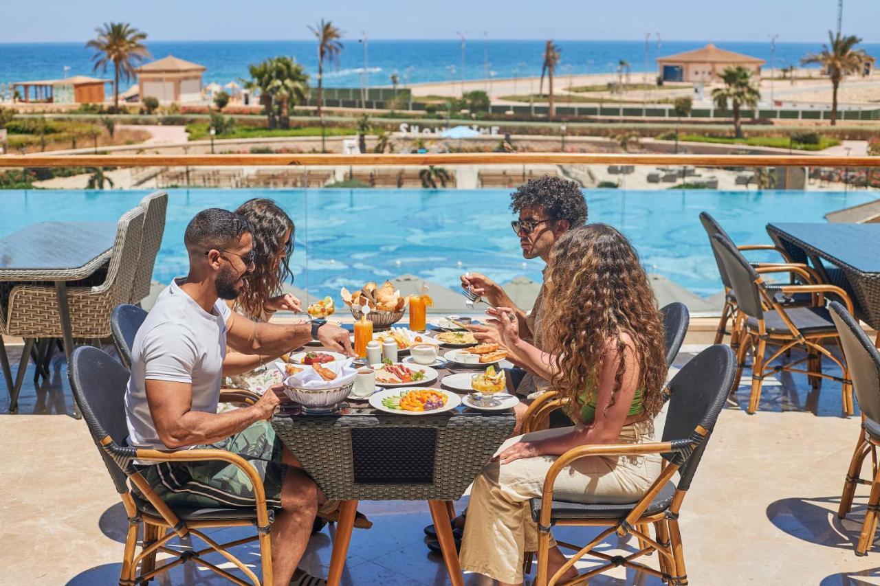 Tolip Resort El Galala Majestic Ain Suchna Exterior foto The photo shows a group of five people enjoying a meal together outdoors, likely at a restaurant or cafe. They are seated at a table with a beautiful view of a swimming pool and the sea in the background. The table is filled with various dishes, and 