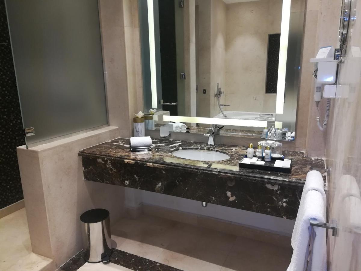 Tolip Resort El Galala Majestic Ain Suchna Exterior foto The photo shows a modern bathroom with elegant design features. There is a large vanity made of dark marble with a sink and a set of toiletries neatly arranged on top. A mirror with built-in lighting is mounted above the sink. To the side, there is a